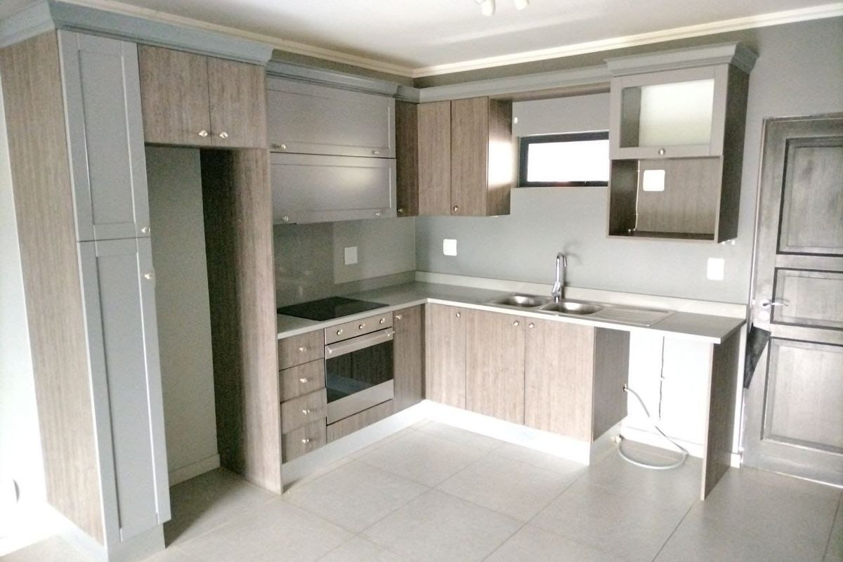 A modern, empty kitchen featuring wood-tone and grey cabinetry, integrated oven and hob, stainless steel sink, and neutral tiled flooring.