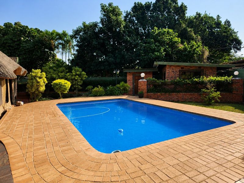 A well-maintained swimming pool area featuring brick paving, a surrounding garden, and a brick outbuilding, set against a backdrop of mature trees.