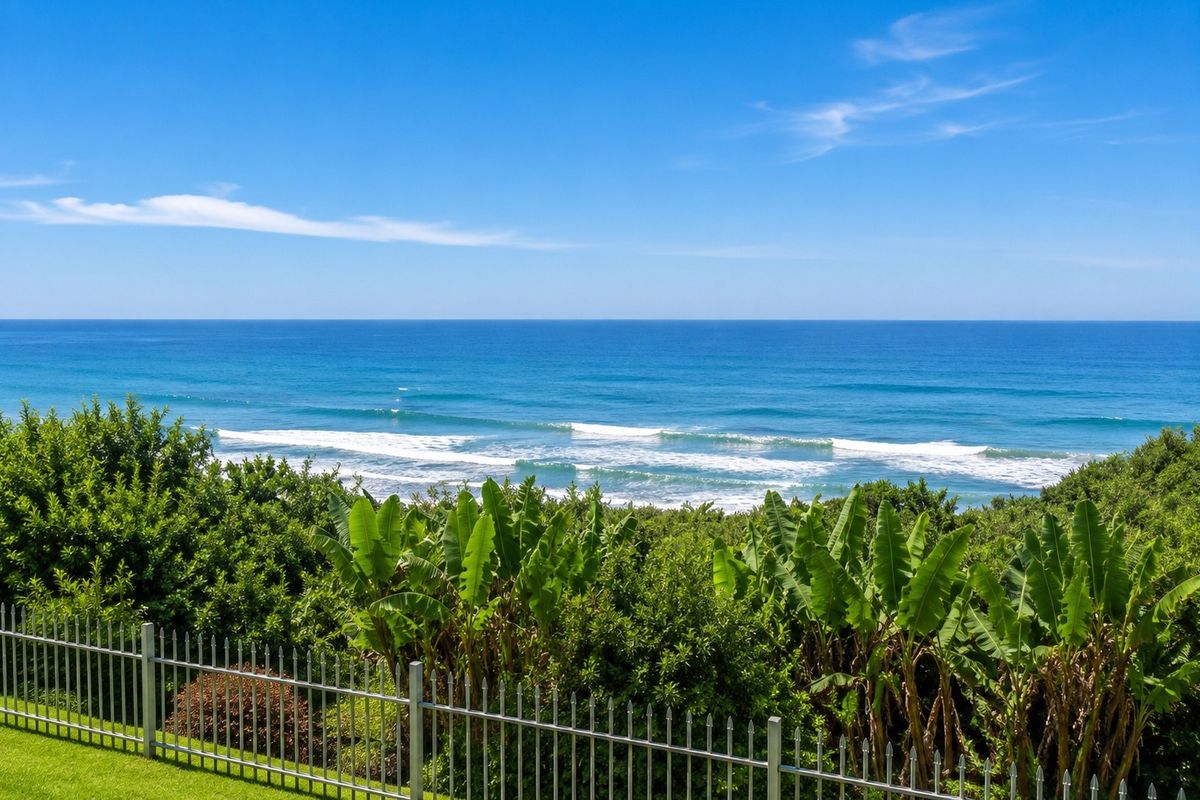 A scenic elevated view of the ocean and coastline, featuring lush greenery and a secure metal perimeter fence.