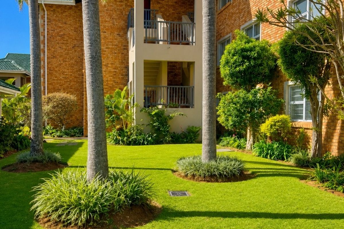 A well-maintained apartment complex exterior featuring brick facade, private balconies, and a lush, landscaped garden area with mature palm trees.