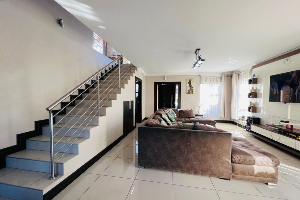 Spacious open-plan living area featuring modern tiled flooring, a sleek staircase with stainless steel balustrades, and a built-in media wall.