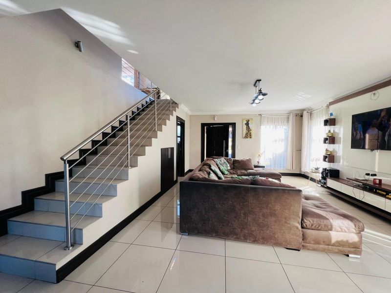 Spacious open-plan living area featuring modern tiled flooring, a sleek staircase with stainless steel balustrades, and a built-in media wall.