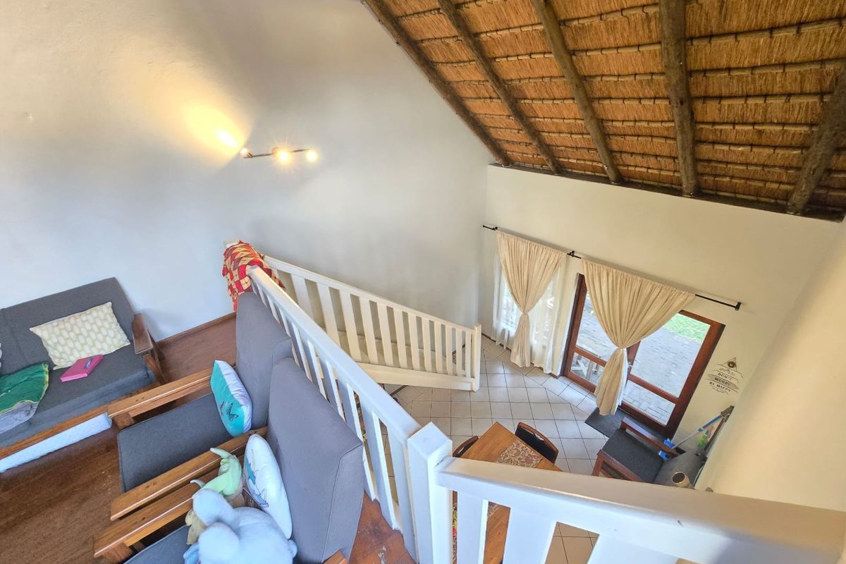 A double-volume living area featuring a traditional thatched roof with exposed rafters, timber staircase, and direct access to the outdoor area via sliding doors.