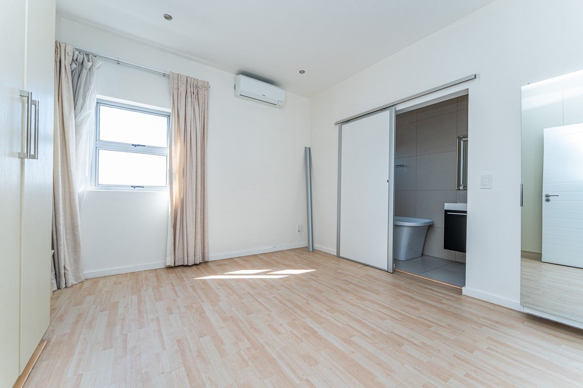 A bright, empty bedroom featuring laminate flooring, a built-in wardrobe, an air conditioning unit, and an en-suite bathroom accessed via a sliding door.