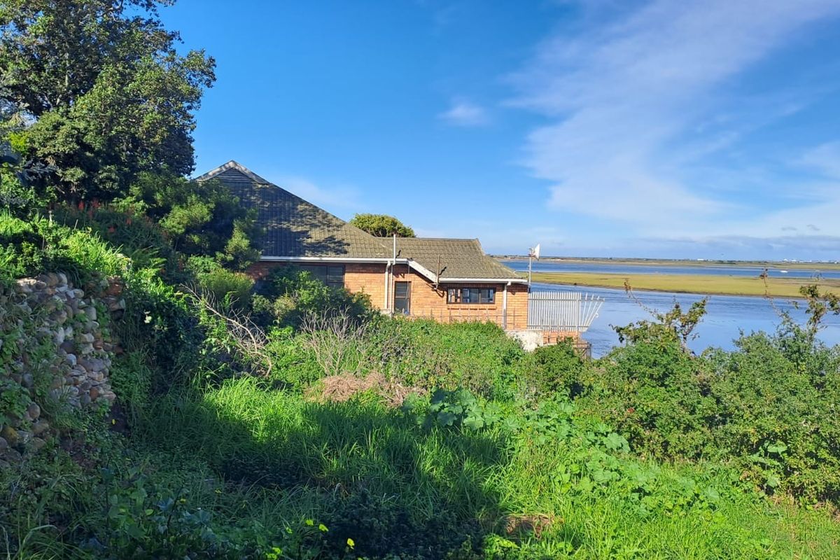 Rear view of a brick residential property situated along a waterfront, featuring a tiled roof and metal balcony railing, surrounded by dense vegetation.