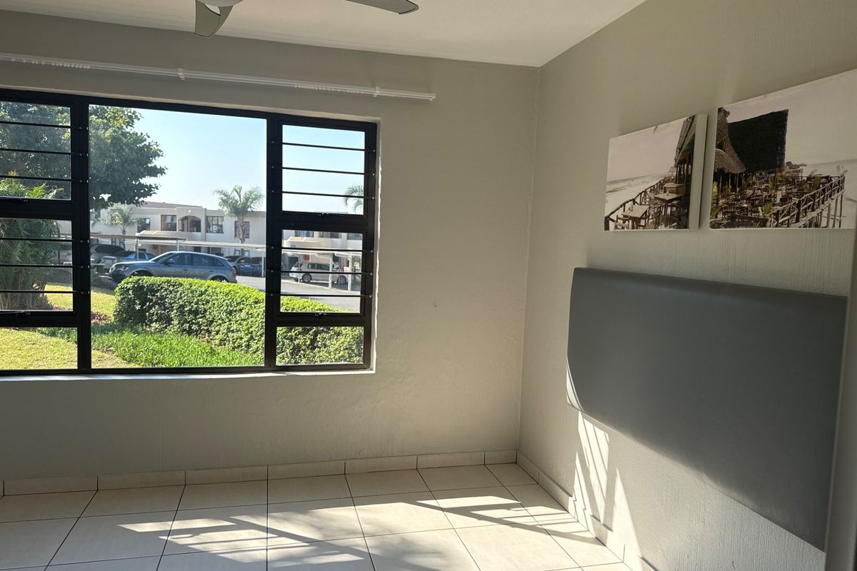A bright, empty bedroom featuring tiled flooring, a ceiling fan, and large windows with security bars overlooking the complex grounds.