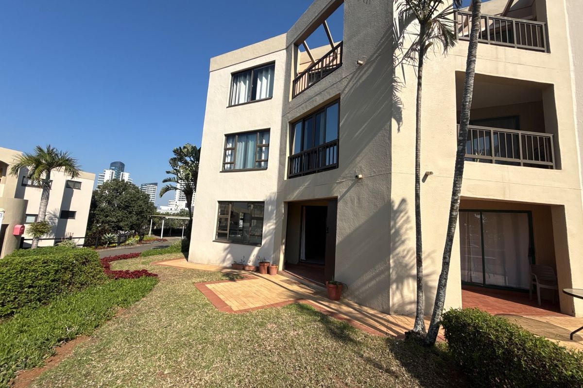 Rear view of a multi-story apartment building featuring private balconies, sliding glass doors, and a paved patio area overlooking a landscaped garden.