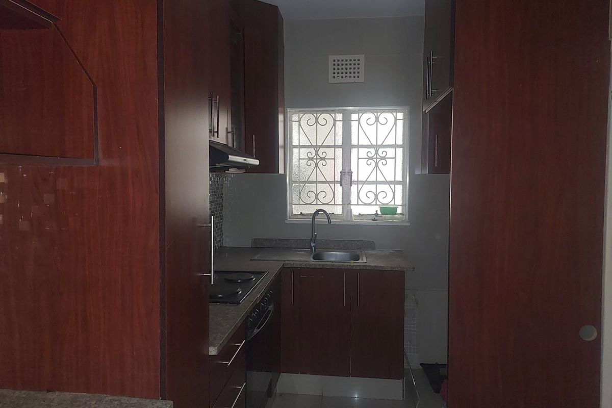 A compact kitchen featuring dark wood-finish cabinetry, laminate countertops, a built-in electric hob, and a window with security bars.