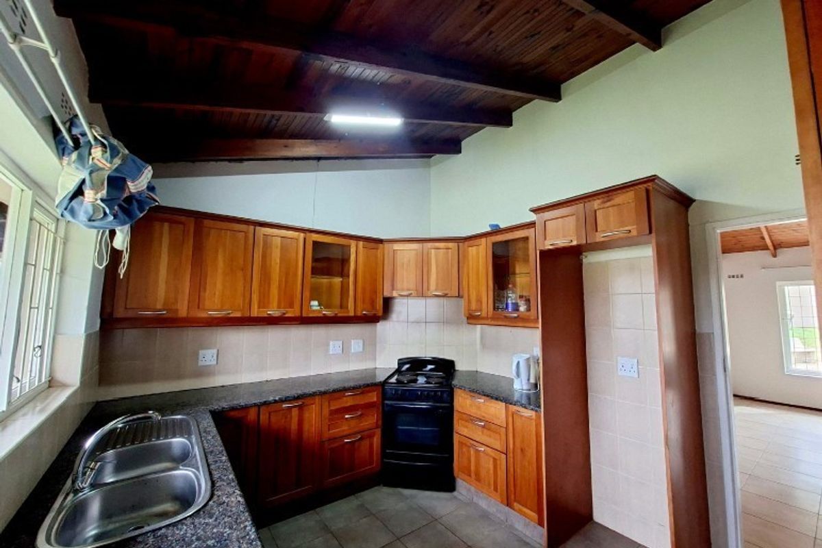 A functional kitchen featuring wooden cabinetry, granite countertops, a double stainless steel sink, and exposed timber beam ceilings with tiled flooring.