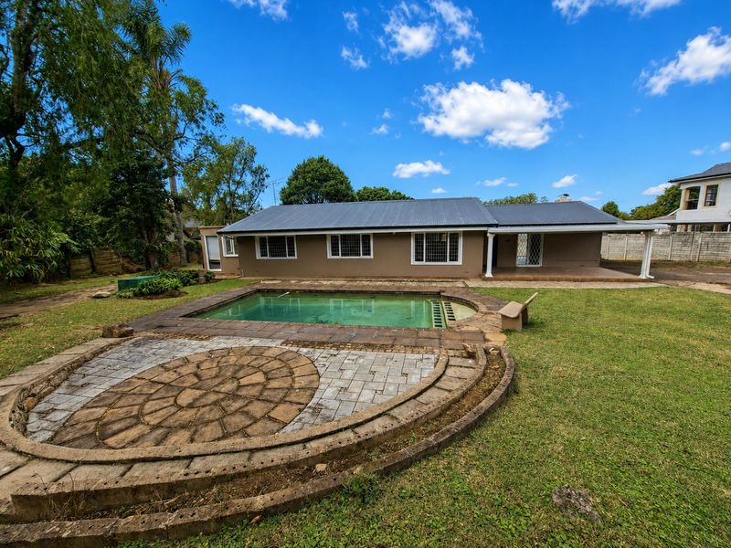 Rear view of the property featuring a swimming pool, circular paved patio area, covered veranda, and a spacious garden with a metal roof structure.