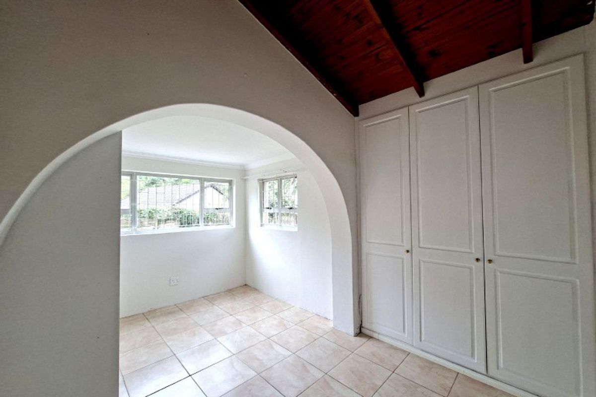 A bright, empty bedroom featuring tiled flooring, built-in cupboards, and a distinctive arched transition with exposed timber ceiling beams.