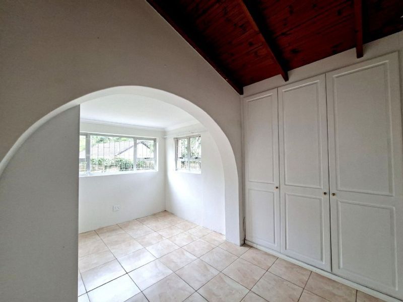A bright, empty bedroom featuring tiled flooring, built-in cupboards, and a distinctive arched transition with exposed timber ceiling beams.