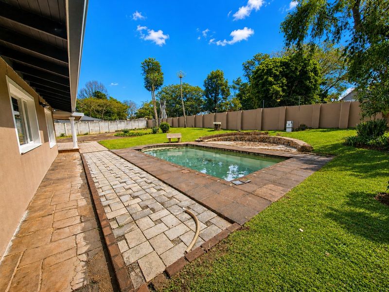 A spacious backyard featuring a swimming pool with a stone retaining wall, surrounded by a paved patio and a well-maintained lawn.