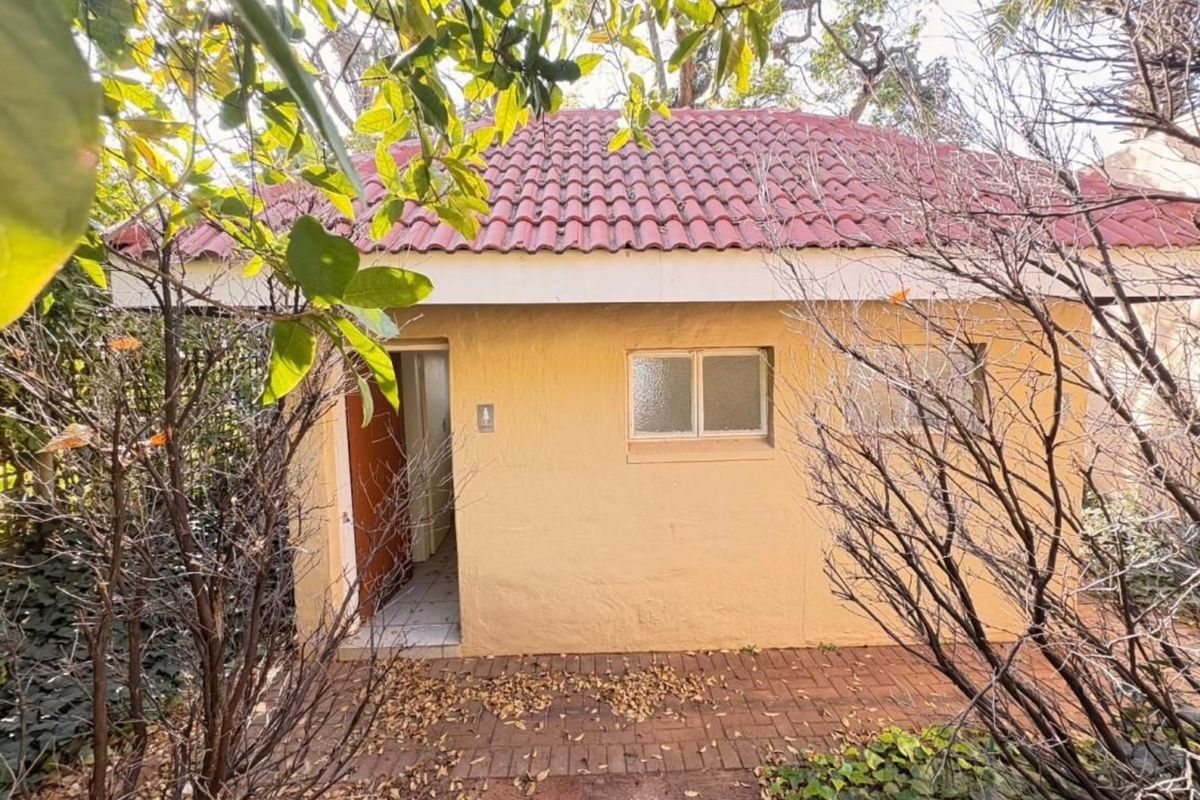 A rear exterior view of a small outbuilding featuring a terracotta tiled roof, brick paved patio area, and mature surrounding garden foliage.