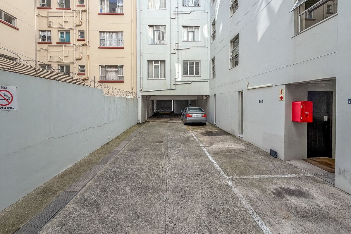 Rear exterior view of the apartment building showing a concrete driveway, parking area, and wall-mounted fire safety equipment.