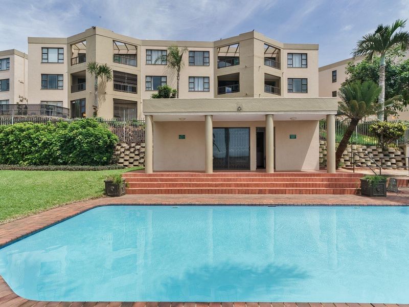 A well-maintained communal swimming pool area featuring brick paving, a covered patio with concrete columns, and landscaped garden surroundings within an apartment complex.