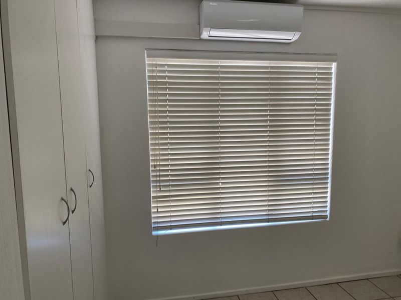 A clean, neutral bedroom featuring built-in cupboards, tiled flooring, and a wall-mounted split-system air conditioner.