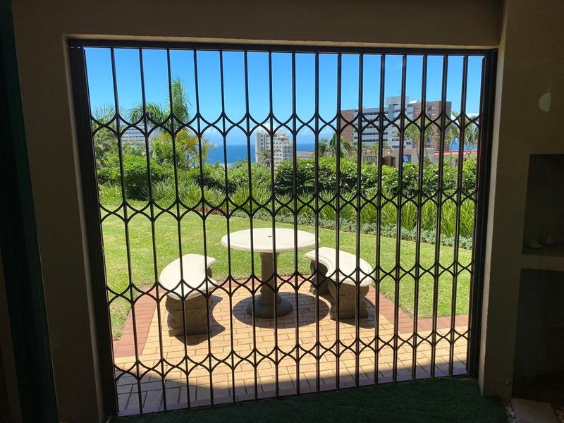 Covered patio area featuring a security gate, paved flooring, and a built-in braai, overlooking a private garden with ocean and city views.