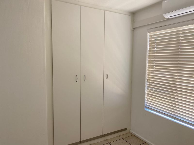A clean bedroom featuring a built-in white wardrobe, tiled flooring, venetian blinds, and a wall-mounted air conditioning unit.