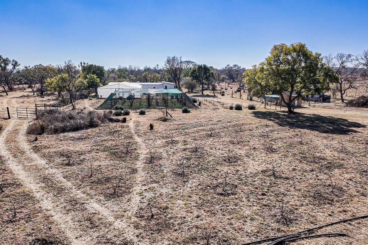 Aerial view of the farm property featuring open land, established trees, agricultural netting structures, and outbuildings under a clear sky.