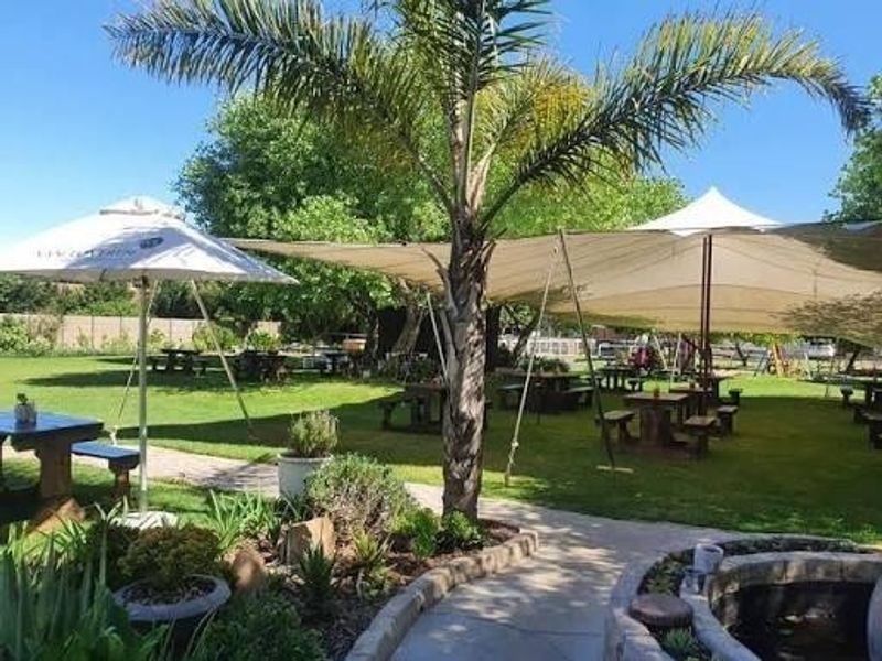 A well-maintained garden area featuring a paved walkway, a stone-edged water feature, mature landscaping, and large tension shade sails for outdoor seating.