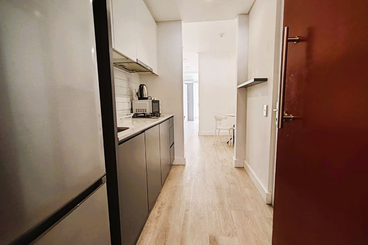 A modern galley-style kitchen featuring sleek cabinetry, a subway tile backsplash, and light wood-look laminate flooring, leading into an open-plan living area.