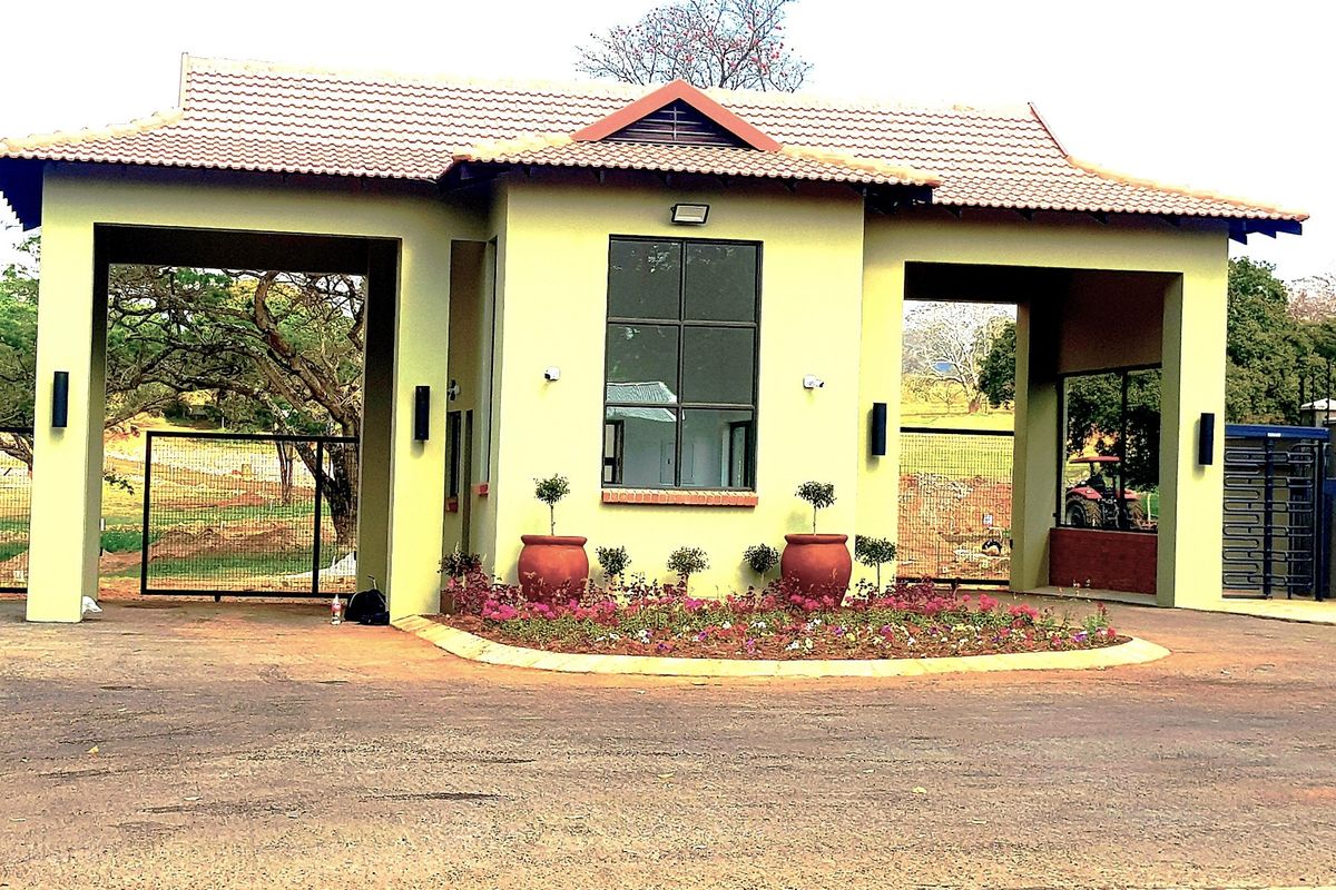 A well-maintained residential security gatehouse featuring a tiled roof, paved driveway, and landscaped garden beds at the entrance.
