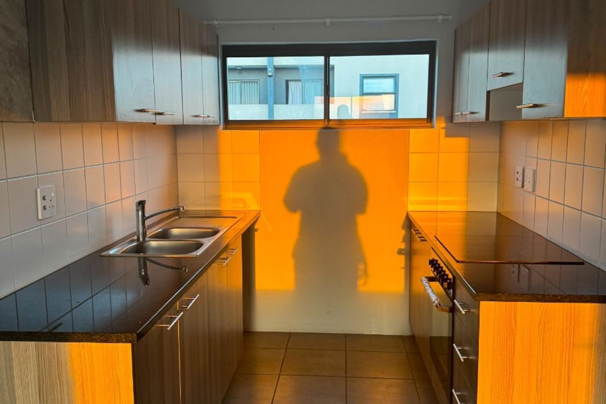 A functional galley-style kitchen featuring wood-finish cabinetry, dark countertops, a double sink, and built-in oven, illuminated by warm natural light.