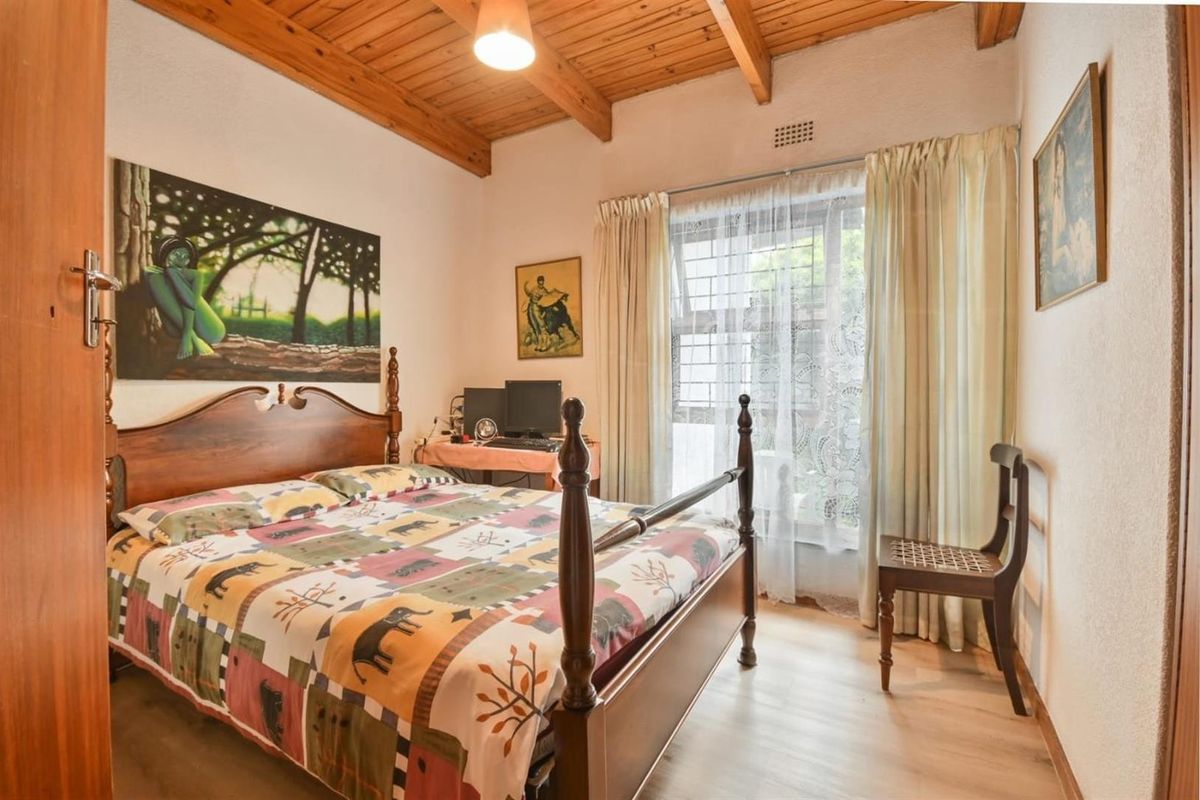 A cozy bedroom featuring exposed timber ceiling beams, laminate flooring, and a window fitted with security burglar bars.