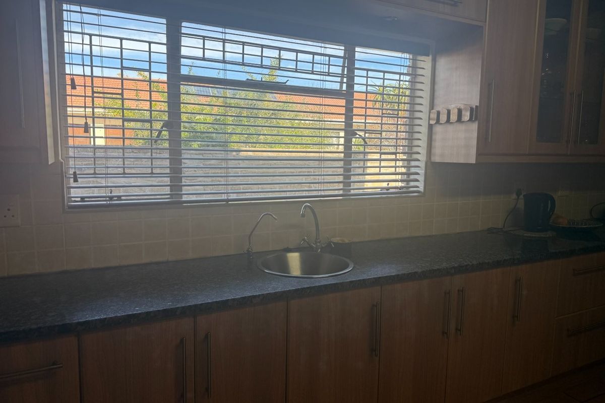 A functional kitchen area featuring wood-look laminate cabinetry, granite countertops, a stainless steel sink, and a tiled backsplash with natural light.