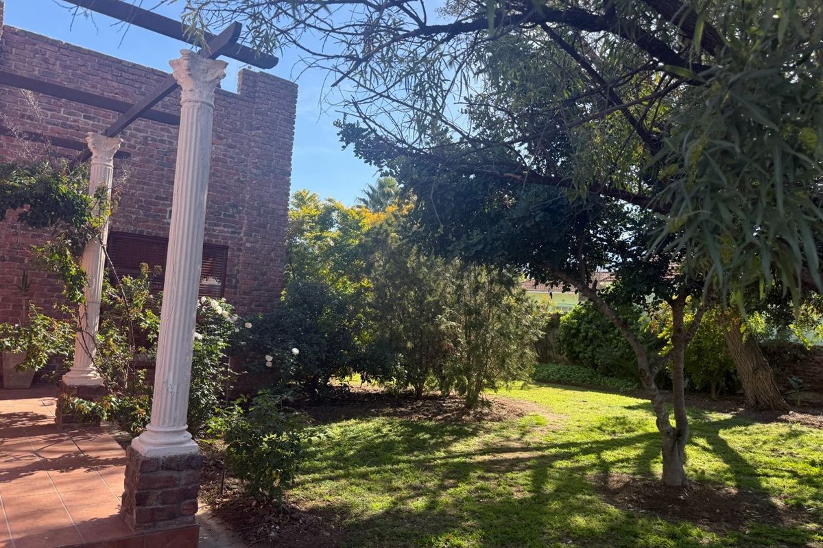 A rear garden view featuring a tiled patio with classical columns, an exposed brick facade, and mature trees providing shade over the lawn.