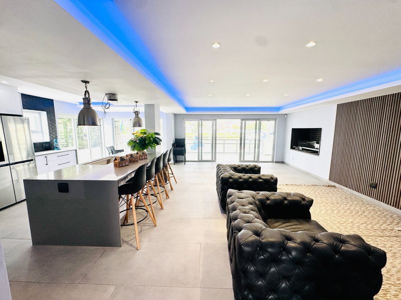 Modern open-plan living area featuring a large kitchen island, sleek tiled flooring, recessed LED lighting, and a feature slatted wood wall with built-in fireplace.