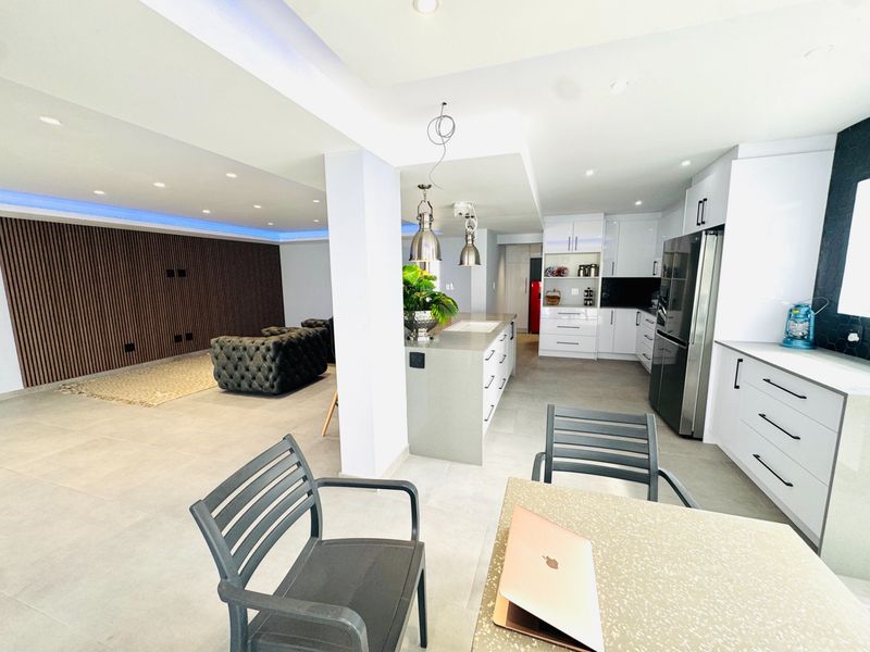 Modern open-plan living and kitchen area featuring sleek white cabinetry, a central island, and contemporary slatted wood wall detailing with ambient cove lighting.