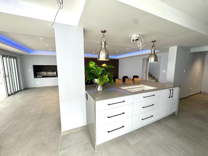 Modern open-plan living area featuring a sleek kitchen island with stone countertops, recessed LED lighting, and a built-in indoor braai area.