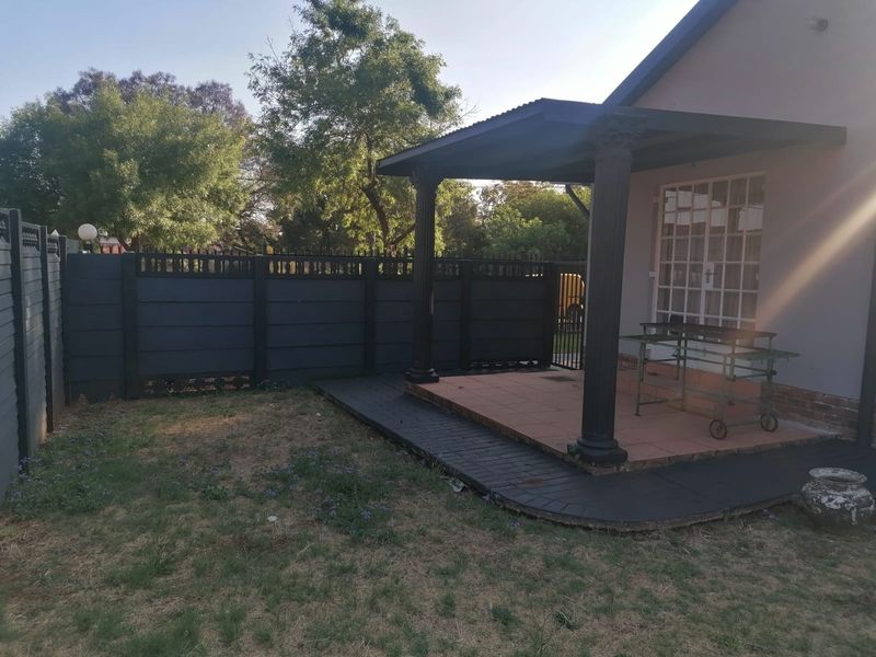 Rear exterior view featuring a covered patio with decorative columns, tiled flooring, and a paved border, set within a fenced garden area.