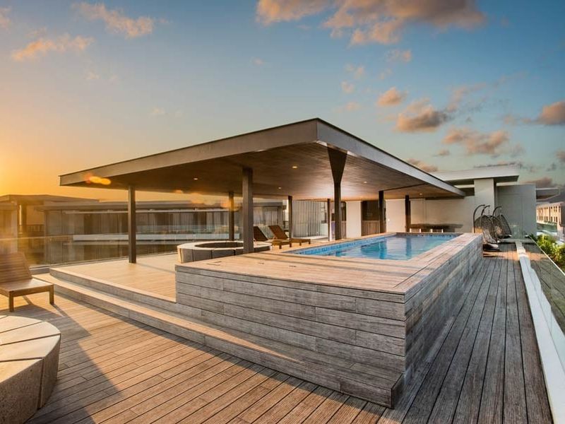 A modern rooftop terrace featuring a raised swimming pool, extensive timber decking, and a covered outdoor lounge area with glass balustrades.