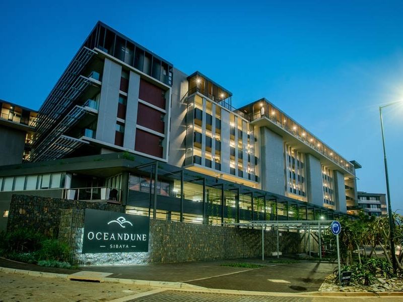Modern multi-story apartment complex exterior featuring contemporary architectural design, stone cladding, and illuminated signage at the entrance.