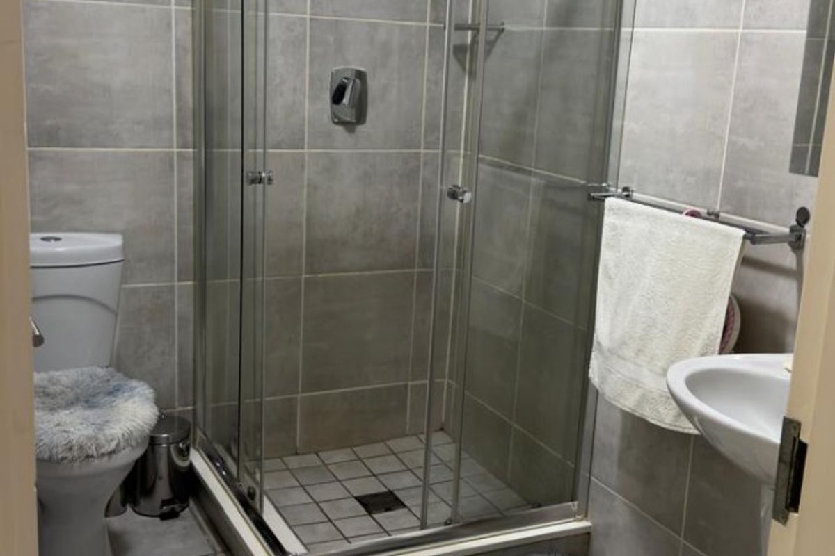 A functional bathroom featuring a glass shower enclosure, pedestal basin, and toilet with neutral grey wall and floor tiling.