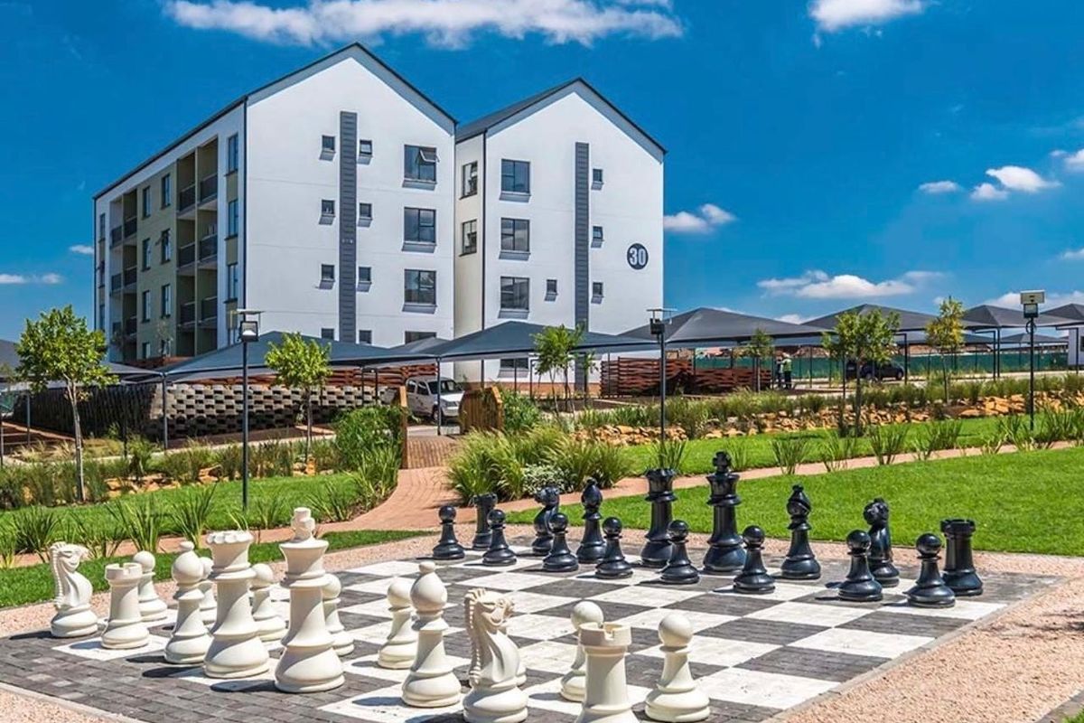 Exterior view of an apartment complex featuring a landscaped garden with a large outdoor chess board and covered parking facilities.