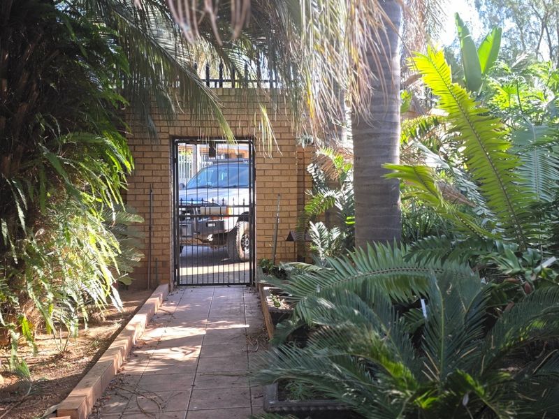 A private garden pathway leading to a brick-walled entrance with a secure metal gate, surrounded by established tropical foliage.
