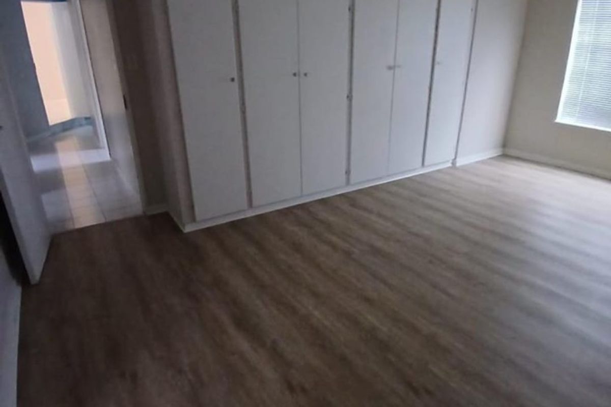 A spacious, empty bedroom featuring neutral laminate flooring, built-in floor-to-ceiling cupboards, and natural light through a window with fitted blinds.