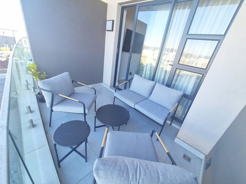 A modern apartment balcony featuring tiled flooring, a sleek glass balustrade, and large sliding glass doors providing access to the interior living space.