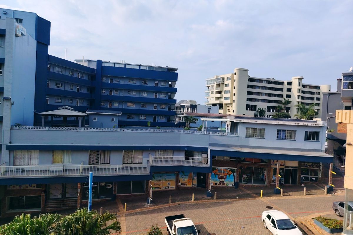 An elevated view of a multi-story apartment complex featuring a paved courtyard, commercial storefronts on the ground level, and residential balconies above.