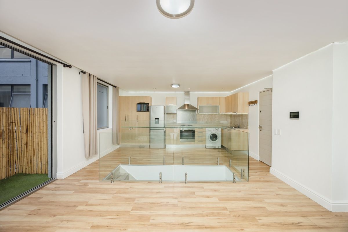 Modern open-plan kitchen and living area featuring light wood cabinetry, integrated appliances, laminate flooring, and a glass balustrade overlooking a lower level.
