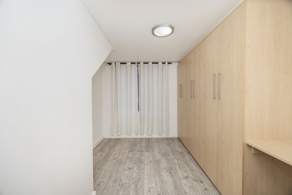 A clean, empty bedroom featuring light wood-tone built-in cupboards, modern laminate flooring, and a window with neutral curtains.