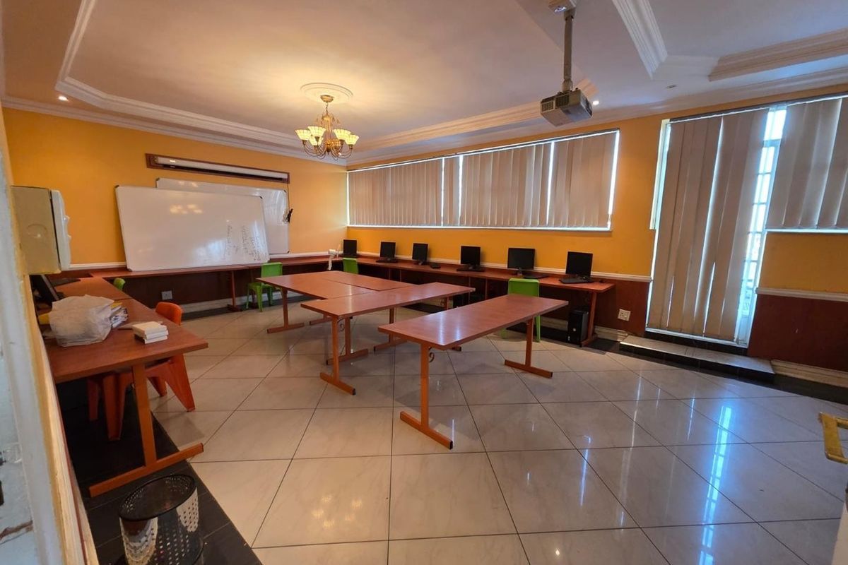 A functional office space featuring tiled flooring, built-in desk workstations, vertical blinds, and a ceiling-mounted projector, suitable for a classroom or training environment.