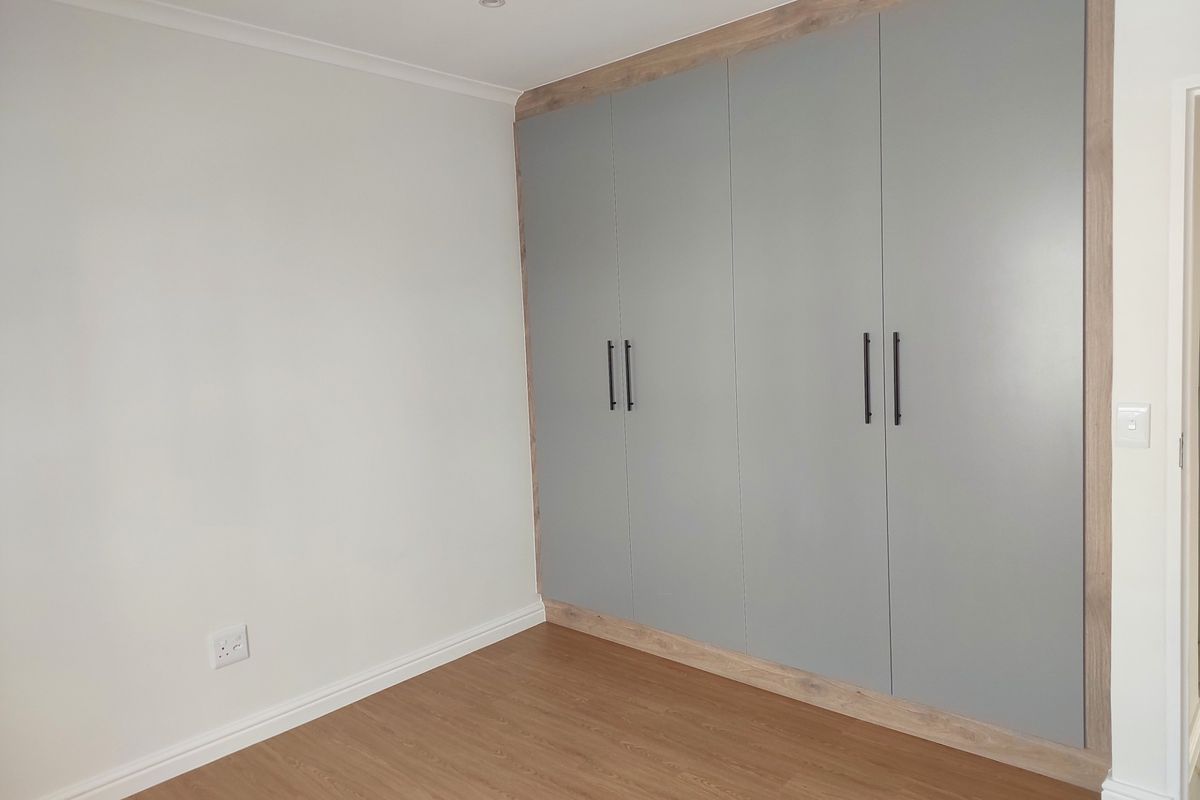 A clean, modern bedroom featuring neutral walls, light wood-look laminate flooring, and floor-to-ceiling built-in cupboards with contemporary grey doors and timber-look framing.