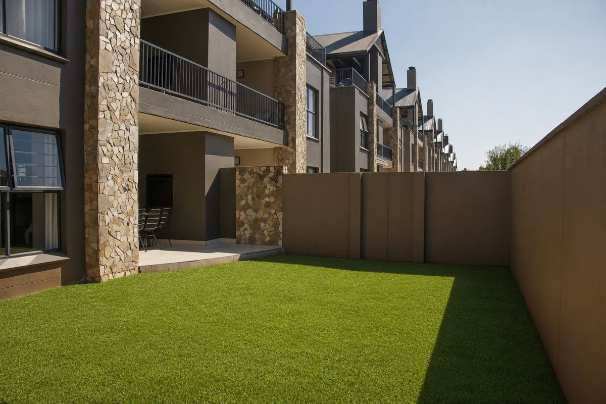 A well-maintained rear exterior view featuring a private garden with artificial lawn, a covered patio, and stone-clad architectural detailing.