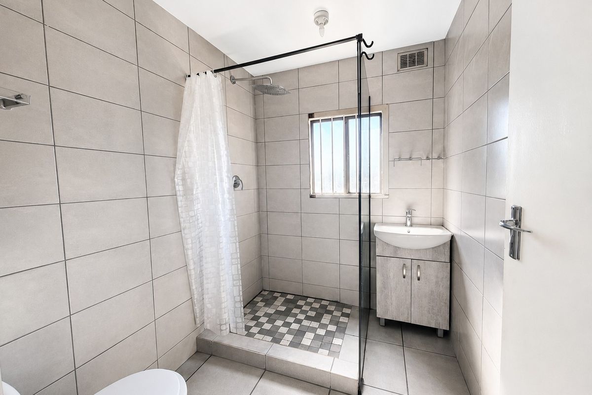 A clean, modern bathroom featuring a walk-in shower with a glass screen, a vanity unit with a basin, and neutral floor-to-ceiling wall tiles.