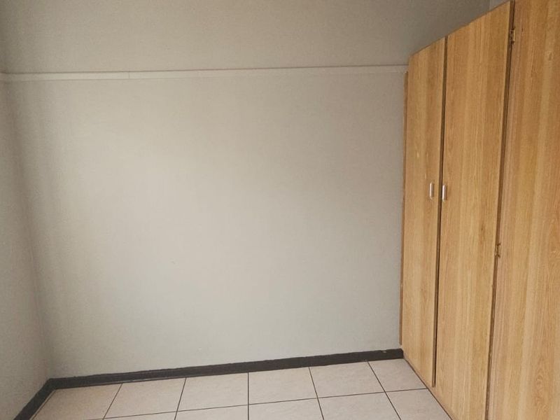 A clean, empty bedroom featuring light-toned built-in cupboards, tiled flooring, and a window with security bars.